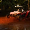 A buggy powers through a muddy pool on a shaded jungle track, throwing up a wall of spray