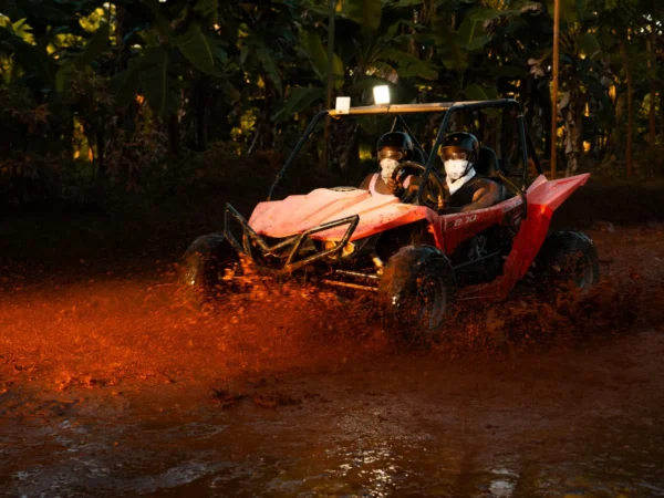 A buggy powers through a muddy pool on a shaded jungle track, throwing up a wall of spray