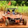 An orange buggy splashes along a wet track past open fields, both riders in helmets and face scarves