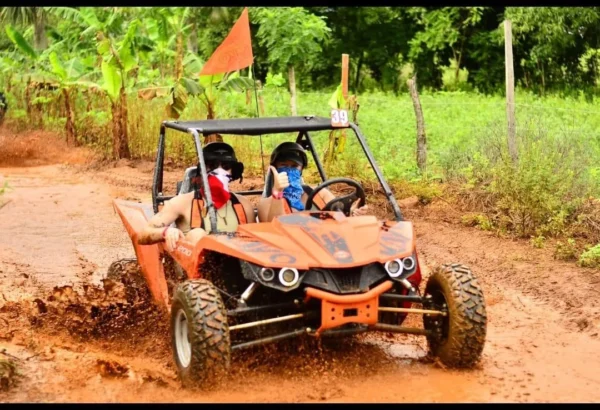 An orange buggy splashes along a wet track past open fields, both riders in helmets and face scarves