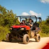 Two riders wave from a red side-by-side buggy on a sandy track under a blue sky