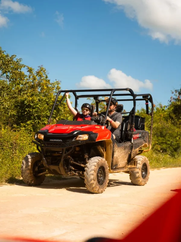 Two riders wave from a red side-by-side buggy on a sandy track under a blue sky