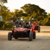 Two riders in helmets sit in red off-road buggies on a sandy track in evening light