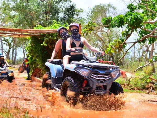 Two riders on a quad bike cross an orange mud puddle as other guests wait behind