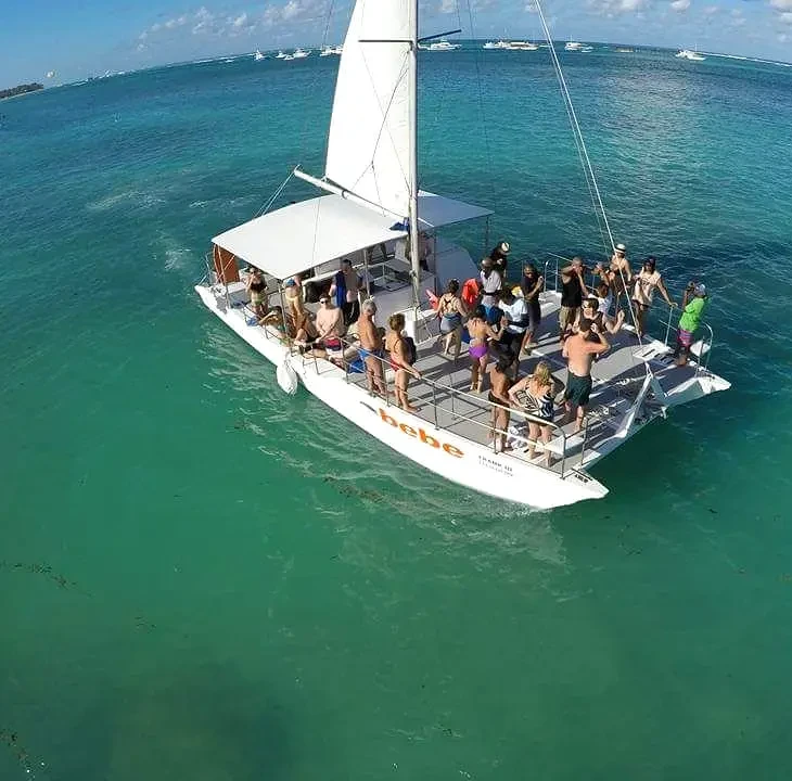 Guests fill the deck of a catamaran under sail on clear turquoise water