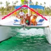 A group of guests sits on the bow netting beneath a decorated arch, resorts lining the shore behind
