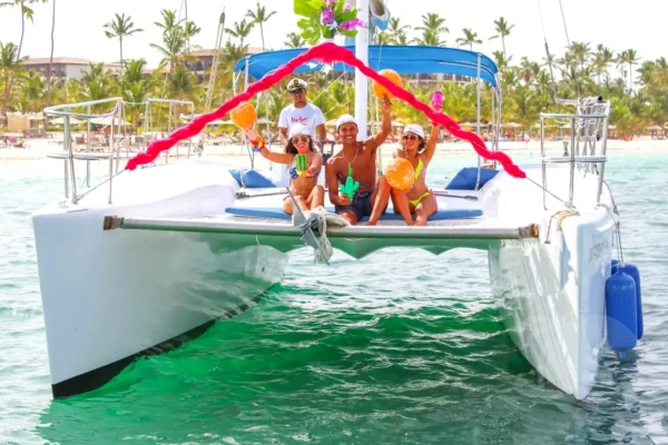 A group of guests sits on the bow netting beneath a decorated arch, resorts lining the shore behind