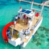 Guests stand on the deck of a moored power catamaran with a red slide, above clear shallow water