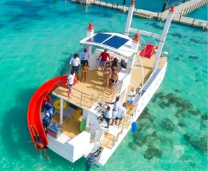 Guests stand on the deck of a moored power catamaran with a red slide, above clear shallow water