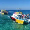 Guests line the rail of a yellow party boat as a second crowded boat follows across open water