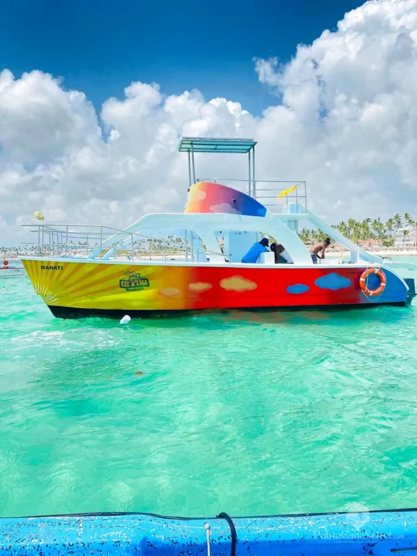 A rainbow-painted party catamaran sits in shallow turquoise water off a resort shoreline