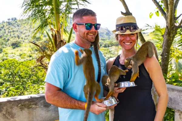 A couple laugh as squirrel monkeys climb across them with green hills behind