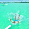 A guest landing backwards in a burst of spray after jumping from the boat slide, with the palm-lined coastline and other boats behind