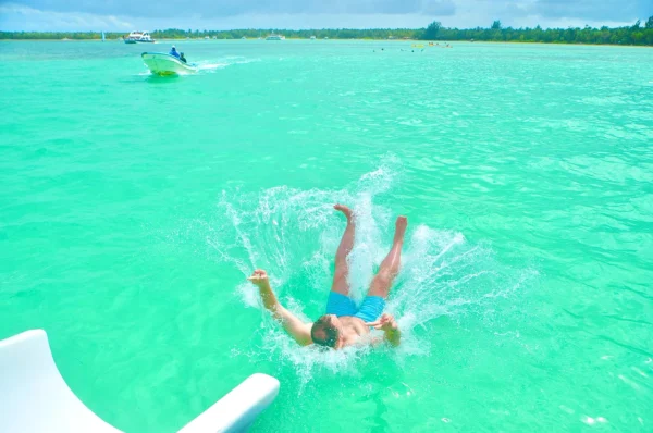 A guest landing backwards in a burst of spray after jumping from the boat slide, with the palm-lined coastline and other boats behind