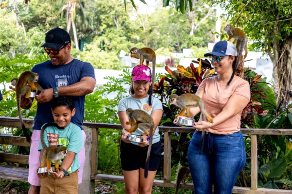 A family stands at a wooden rail with squirrel monkeys perched on their shoulders and arms, bright croton leaves behind