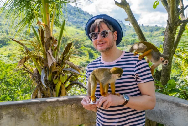 A guest in a striped shirt holds a feeding cup while two squirrel monkeys climb his arm and shoulder