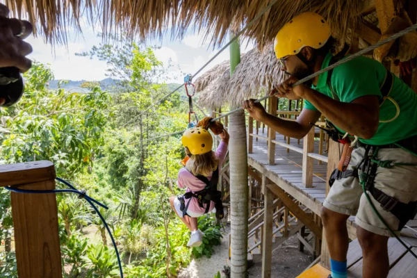 Child launching from the zipline platform with a guide A child in a yellow helmet sets off along a zipline as a guide steadies the cable from the platform
