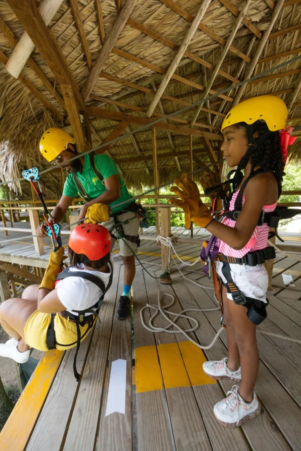 Guide fitting safety harnesses in the zipline hut A guide fits safety harnesses on two young guests under the thatched roof of the launch hut