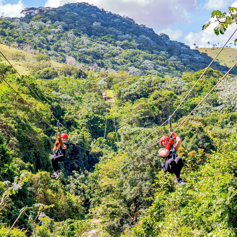 Two guests in red helmets ride parallel zipline cables above the jungle canopy