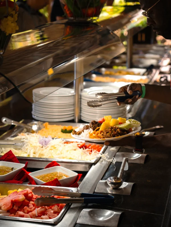 A buffet line of Dominican dishes with a guest lifting a plate of rice, stewed meat and fresh fruit